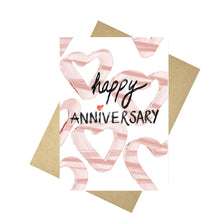 Load image into Gallery viewer, White card featuring simple heart shapes in a mix of pale and mid pink under the words happy anniversary in black textured handwriting with a red love heart for the i in anniversary. Behind the card a brown envelope is visible, behind which is a white background.
