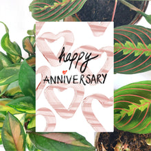 Load image into Gallery viewer, White card featuring simple heart shapes in a mix of pale and mid pink under the words happy anniversary in black textured handwriting with a red love heart for the i in anniversary. Behind the card a brown envelope is visible, in front of a collection of houseplants on a white background.
