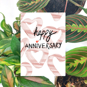 White card featuring simple heart shapes in a mix of pale and mid pink under the words happy anniversary in black textured handwriting with a red love heart for the i in anniversary. Behind the card a brown envelope is visible, in front of a collection of houseplants on a white background.