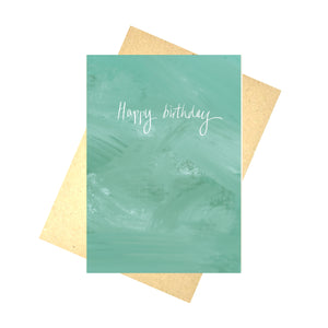 A turquoise tonal card sits in front of a recycled brown paper envelope, in front of a white background. The card features the words 'Happy birthday' in white handwriting.