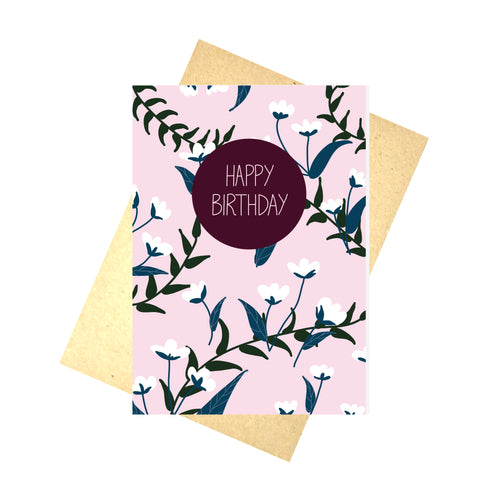 Pale pink card featuring an all over blue and white floral pattern with green vines. Towards the top centre of the card is a plum coloured circle with the words 'HAPPY BIRTHDAY' in pale pink. Underneath the card you can see a recycled brown paper envelope which is on top of a white background.