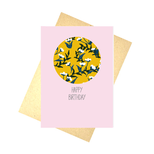 Pale pink card featuring a mustard yellow circle with a simple floral pattern. Underneath the circle are the words HAPPY BIRTHDAY in dark grey handwriting. Behind the card is a brown recycled paper envelope, behind which is a white background.