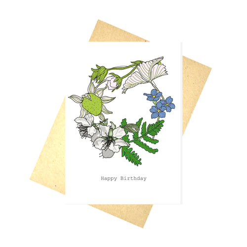 White card featuring a wreath design made up of different wild plants, including a strawberry and forgetmenots. Below the wreath are the words 'Happy Birthday' are on the card in black. Behind the card is a brown envelope, behind which is a white background. The strawberry is green while it's leaves and stem are a contrasting baby pink, next to it is the warm blue and yellow forgetmenot, below which is a green fern like plant, bordering the blossom.