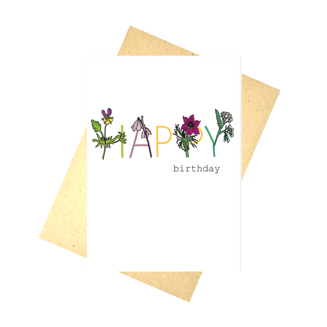 White card featuring the words happy birthday. Happy is written in a mix of colours with flowers making up part of each letter. H is heartsease, A is autumn snowflake, P is pasque flower and Y is yarrow. Behind the card is a brown envelope, behind which is a white background.
