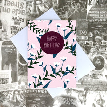 Load image into Gallery viewer, Pale pink card featuring an all over blue and white floral pattern with green vines. Towards the top centre of the card is a plum coloured circle with the words &#39;HAPPY BIRTHDAY&#39; in pale pink. Underneath the card you can see a recycled lilac paper envelope which is on top of a white and black printed collagebackground.
