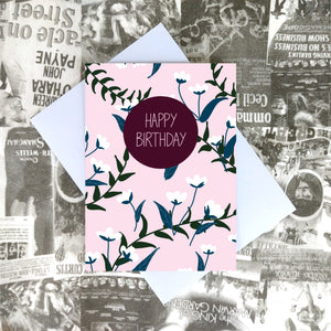 Pale pink card featuring an all over blue and white floral pattern with green vines. Towards the top centre of the card is a plum coloured circle with the words 'HAPPY BIRTHDAY' in pale pink. Underneath the card you can see a recycled lilac paper envelope which is on top of a white and black printed collagebackground.
