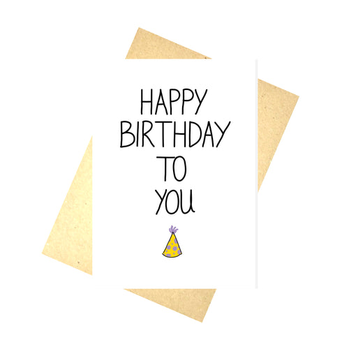 A white card with the words HAPPY BIRTHDAY TO YOU in black writing. Under the words is a little yellow party hat with lilac spots. Behind the card is a brown recycled paper envelope, behind which is a white background.