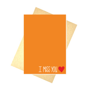 Warm orange card featuring the words 'I MISS YOU' in white in the bottom left next to a small red heart. Behind the card a brown envelope on a white background is visible.