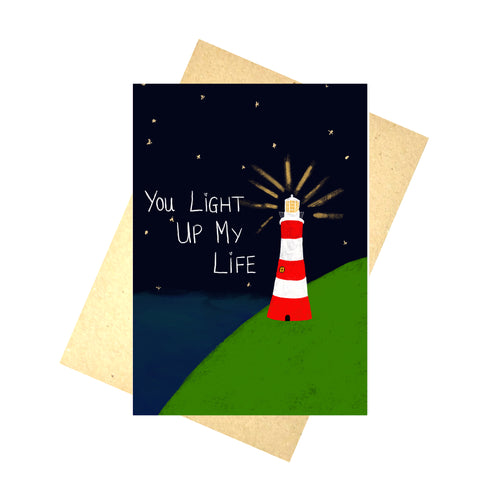 A dark blue card featuring a green hill in front of the sea with a white and red lighthouse lit up in the night sky. The words 'You Light Up My Life' in white handwriting. Behind the card you can see a recycled brown envelope, behind which is a white background.
