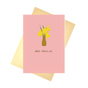 A warm pink card featuring a brown vase filled with yellow daffodils sits above the words 'Happy Mothers Day'. Behind the card is a brown envelope, which is in front of a white background.