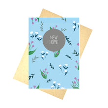 Load image into Gallery viewer, Blue card featuring a simple floral and leaf pattern in greens, white, dark blue and purple. Central and towards the top of the card is a mid grey circle with the words NEW HOME in white in the middle. Behind the card is a brown envelope on top of a white background.
