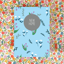 Load image into Gallery viewer, Blue card featuring a simple floral and leaf pattern in greens, white, dark blue and purple. Central and towards the top of the card is a mid grey circle with the words NEW HOME in white in the middle. Behind the card is a floral printed background.
