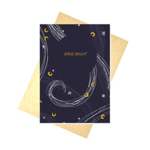 Load image into Gallery viewer, A dark blue card sits on a recycled brown envelope on top of a white background. The card has the words SHINE BRIGHT across it in gold within a circle of dark blue, while around it are stars in white and moons in mustard yellow, as well as some curving lines in white and lighter blue inspired by the wind.
