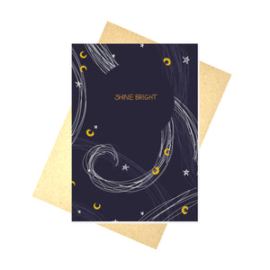 A dark blue card sits on a recycled brown envelope on top of a white background. The card has the words SHINE BRIGHT across it in gold within a circle of dark blue, while around it are stars in white and moons in mustard yellow, as well as some curving lines in white and lighter blue inspired by the wind.