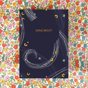 A dark blue card sits on a floral background. The card has the words SHINE BRIGHT across it in gold within a circle of dark blue, while around it are stars in white and moons in mustard yellow, as well as some curving lines in white and lighter blue inspired by the wind. 