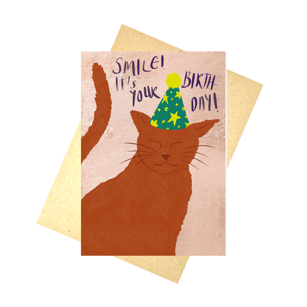 A textured dusky pink card sits in front of a brown envelope on a white background. The card features a ginger cat wearing a bluey green party hat with a yellow pompom and yellow stars and crescent moons on. Above the cat you can see the words 'SMILE! IT'S YOUR BIRTHDAY!' in purple handwriting.