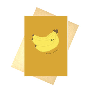 Yellow orange card featuring a cartoon banana bunch with a smiley face. To the bottom right of the bananas are the words Thanks a bunch!! in black writing. Behind the card is a brown envelope, behind which is a white background.