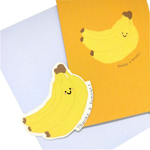 A yellow orange card with a cartoon banana and the words 'Thanks a bunch!!' are to the top right of the pic. Behind the card but slightly further down is a lilac envelope, on top of which is a yellow banana sticker. Behind everything is a white background.