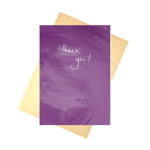 A purple mixed tone card sits on a recycled brown paper envelope in front of a white background. The card features the words 'Thank you!' in messy white handwriting. 