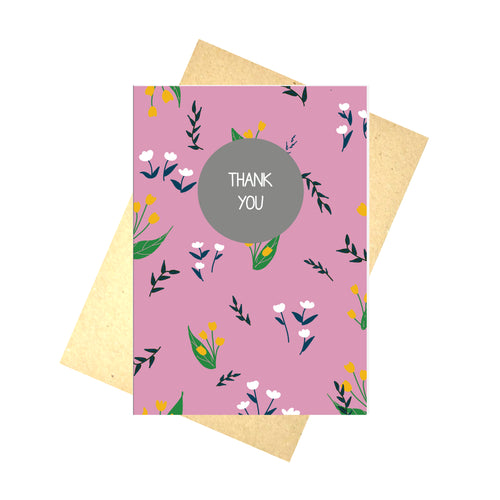 Pink card featuring dark green leafy vines, white fluffy flowers and mustard yellow tulips in an all over pattern. Central, towards the top of the card is a grey circle, with the words THANK YOU written in white. Behind the card is a brown envelope, behind which is a white background.