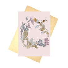 Load image into Gallery viewer, A pale pink card featuring a silhoette wreath made using photos of naturally dyed fabric and a painted background. The wreath is central to the card and features a dashed line circle behind the flowers. Behind the card is a brown envelope, behind which is a white background.
