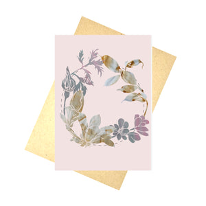 A pale pink card featuring a silhoette wreath made using photos of naturally dyed fabric and a painted background. The wreath is central to the card and features a dashed line circle behind the flowers. Behind the card is a brown envelope, behind which is a white background.