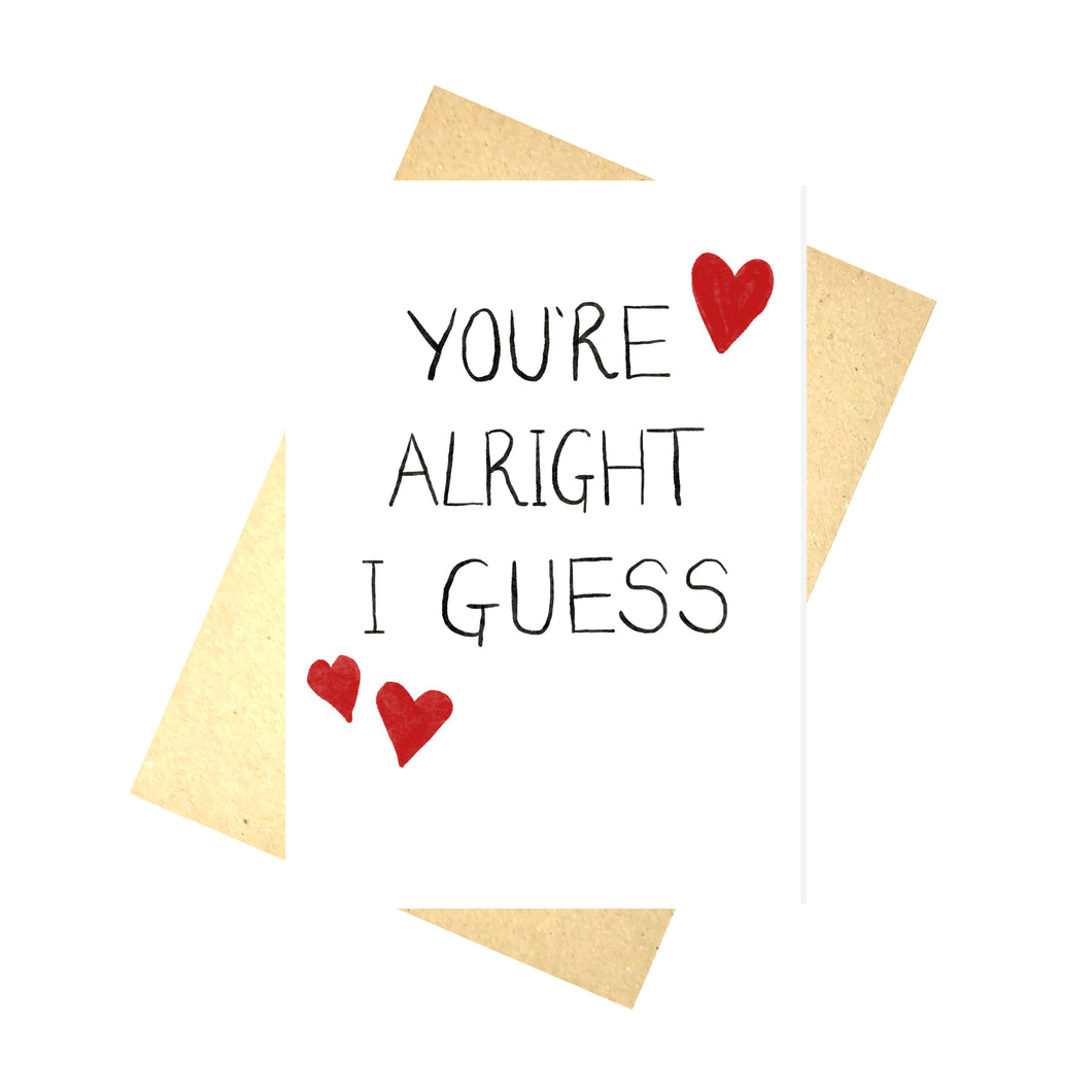 White card with the words ' YOU'RE ALRIGHT I GUESS' in black with red hearts around it. Behind the card is a brown envelope, behind which is a white background.