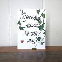Load image into Gallery viewer, A white card featuring the words &#39;You&#39;re Stuck With Me&#39; in the centre. Behind the handwritten words are strands of string of hearts and towards the bottom left of the piece is a red heart. Behind the card is a white wall, and below it is a wooden desk.
