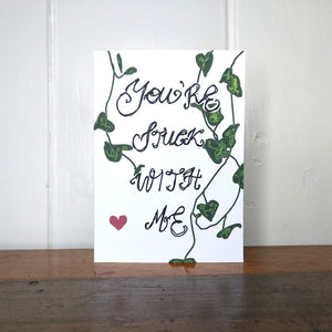 A white card featuring the words 'You're Stuck With Me' in the centre. Behind the handwritten words are strands of string of hearts and towards the bottom left of the piece is a red heart. Behind the card is a white wall, and below it is a wooden desk.