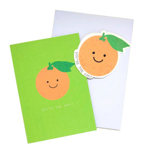 Lime green card featuring a cartoon orange with the words You're the Zest underneath in white writing. Behind the card is a lilac envelope on a white background, in front of the envelope is an orange sticker featuring the same design as the card.