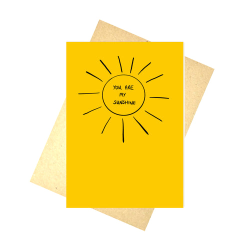 Warm yellow card featuring a simple sun shape in black. In the middle of the sun are the handwritten words 'YOU ARE MY SUNSHINE' in black handwriting. Behind the card you can see a brown envelope, in front of a white background. 