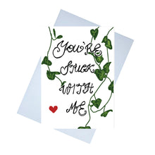 Load image into Gallery viewer, A white card featuring the words &#39;You&#39;re Stuck With Me&#39; in the centre. Behind the handwritten words are strands of string of hearts and towards the bottom left of the piece is a red heart.  Behind the card is a lilac envelope behind which is a white background.
