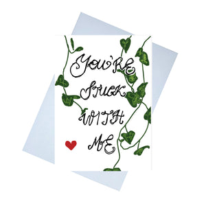 A white card featuring the words 'You're Stuck With Me' in the centre. Behind the handwritten words are strands of string of hearts and towards the bottom left of the piece is a red heart.  Behind the card is a lilac envelope behind which is a white background.